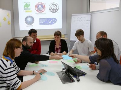 Einige Personen sitzen um einen Tisch mit farbigen Karten und einem Laptop auf dem Tisch, im Hintergrund Logos von Nachhaltigkeitszertifikaten