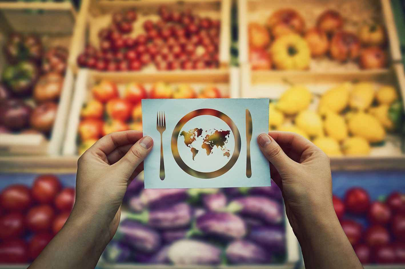 Hände halten ein Blatt Papier, auf dem ein Teller und Besteck zu sehen sind. Auf dem Teller ist eine Weltkarte gezeichnet, im Hintergrund ein Obst- und Gemüseregal im Supermarkt.