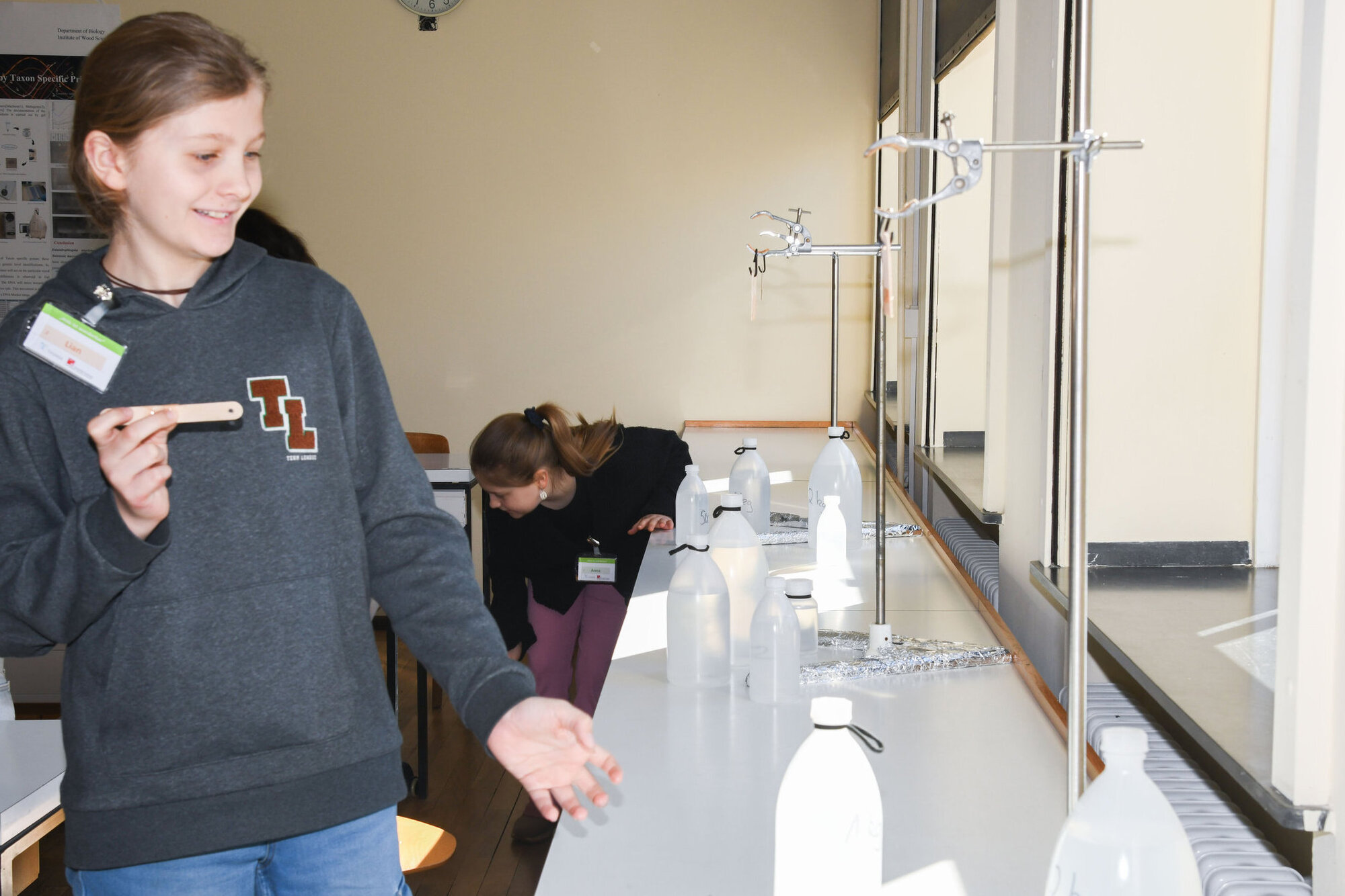 Mädchen in einem Labor, die auf eine Reihe von Flaschen zeigt. Im Hintergrund sind weitere Schülerinnen zu sehen, die an einem Experiment arbeiten.