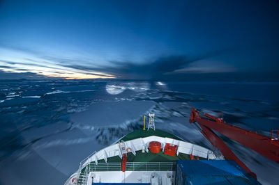 Blick vom Vorderschiff über den Bug eines Schiffes nach vorn. WAsserfläche mit zahlreichen Eisschollen, untergehende Sonne hinter Wolken.