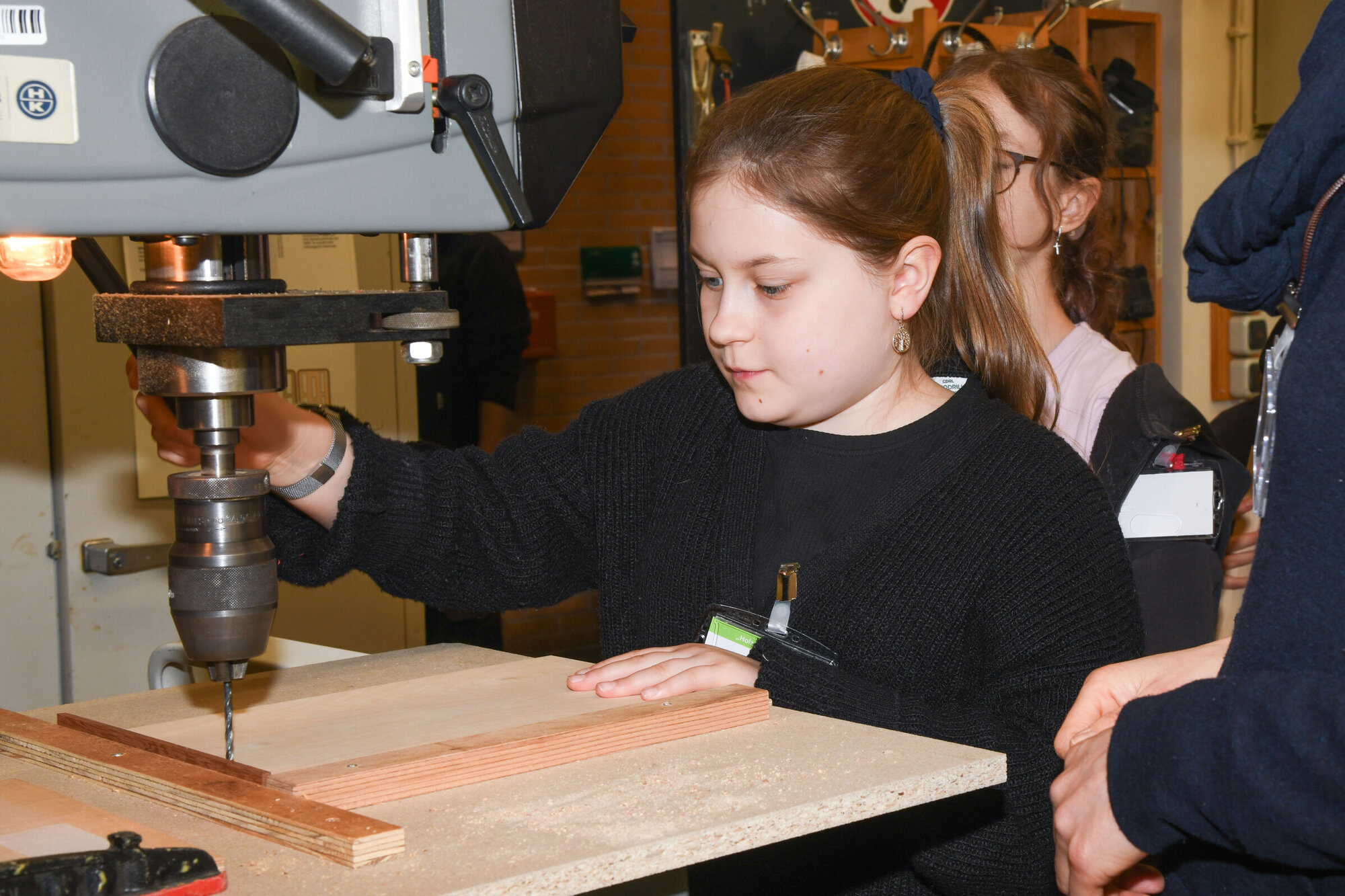 Ein Mädchen in einer Werkstatt vor einer großen Maschine, die ein Loch in ein Holzbrett bohrt.