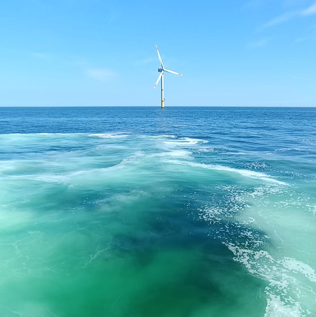 Ein einzelnes Windrad im Meer, im Vordergrund das Schraubenwasser eines Schiffes.