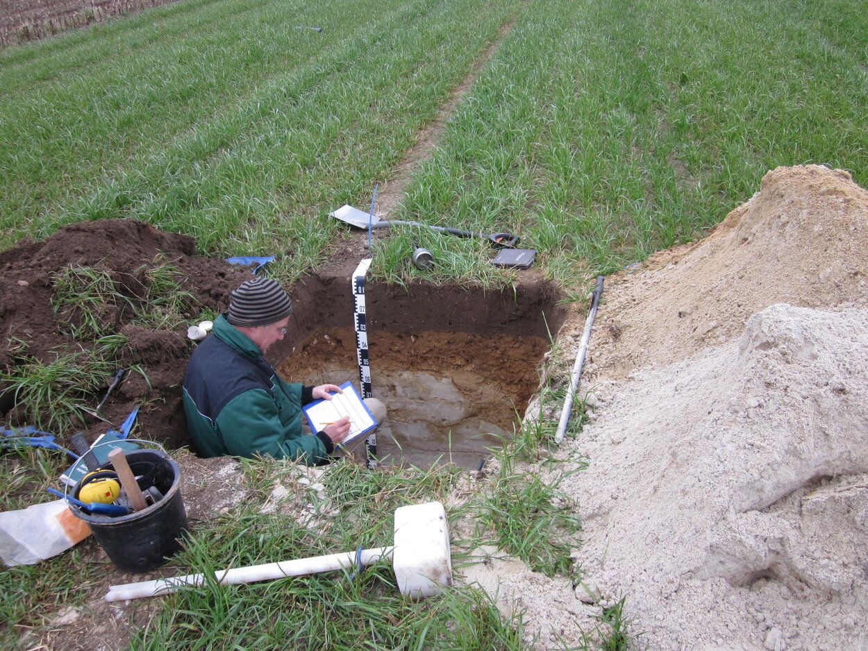 Eine Grube in einem Feld mit Werkzeug auf dem Boden und einer Person in der Grube, die Notizen macht.