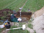 A pit in a field with tools on the ground and a person in the pit taking notes.