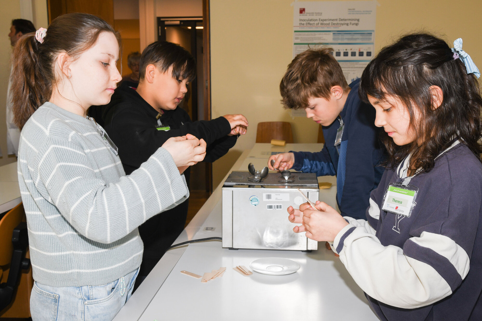 Vier Mädchen und Jungen stehen um einen Tisch herum und hantieren mit kleine Gegenständen. Auf dem Tisch steht eine kompakte Maschine aus Metall. Ein Kind träufelt etwas hinein.