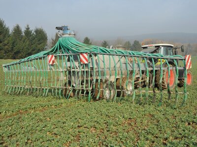 Hinter einem Trecker hängt ein Anhänger mit Gülle, aus dem über Schläuche auf einer breiten Fläche, die Gülle verteilt wird