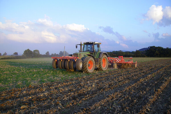 Ein Acker, der mit einem Traktor und landwirtschaftlichem Gerät bearbeitet wird. Im Hintergrund sind Bäume zu sehen, der Himmel ist blau mit ein paar Wolken.