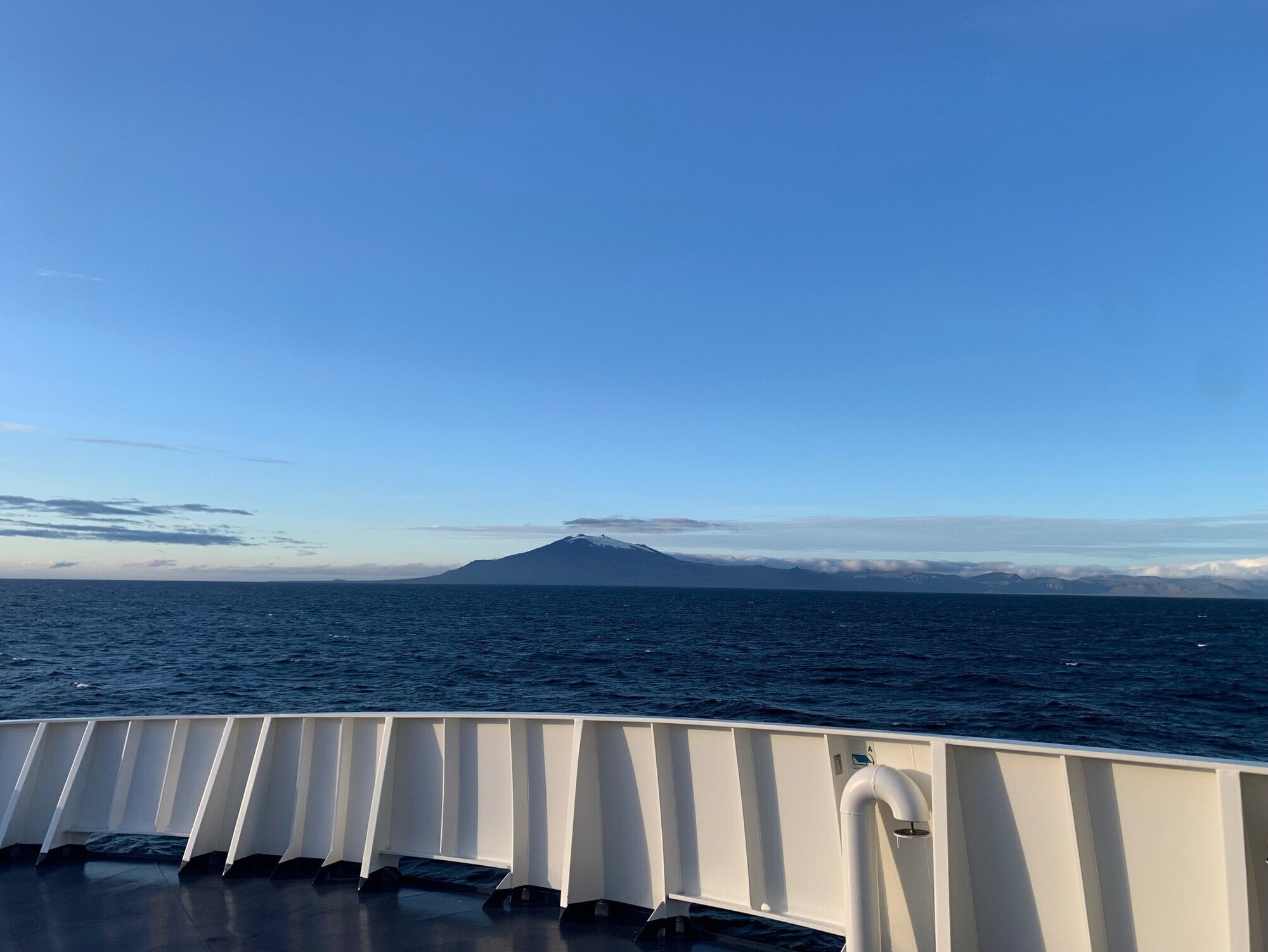 Blick über die Bordwand auf Meer, blauen Himmel und Island in der Ferne.