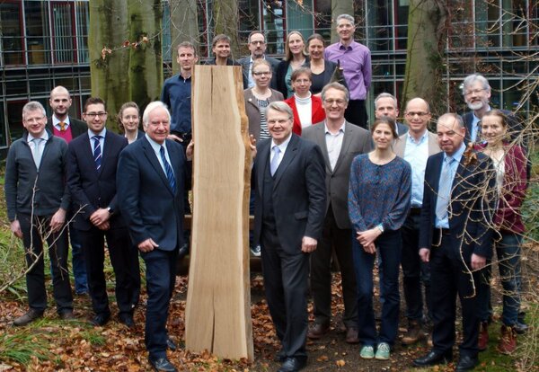 Gruppenfoto vor einem Gebäude, mehrere Menschen stehen um ein großes Holzstück