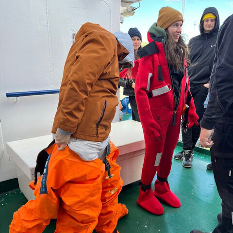 Several people are standing on deck, two of whom are putting on orange/red survival suits.