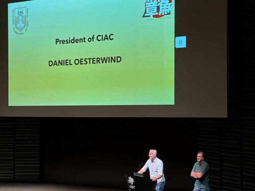 Daniel Oesterwind (am Pult) stellt sich in Okinawa als neuer Präsident des CIAC vor. Eine große Leinwand auf der steht: Persident of CIAC Daniel Oesterwind. Es stehen zwei Männer neben einem Rednerpult.