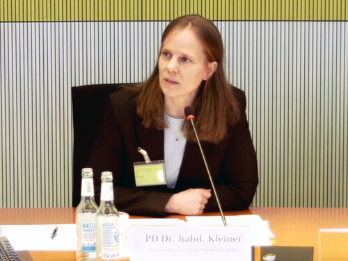 Eine Frau sitzt an einem Holztisch auf einem schwarzen Stuhl und spricht vor einem hellen Hintergrund in ein Mikrofon.