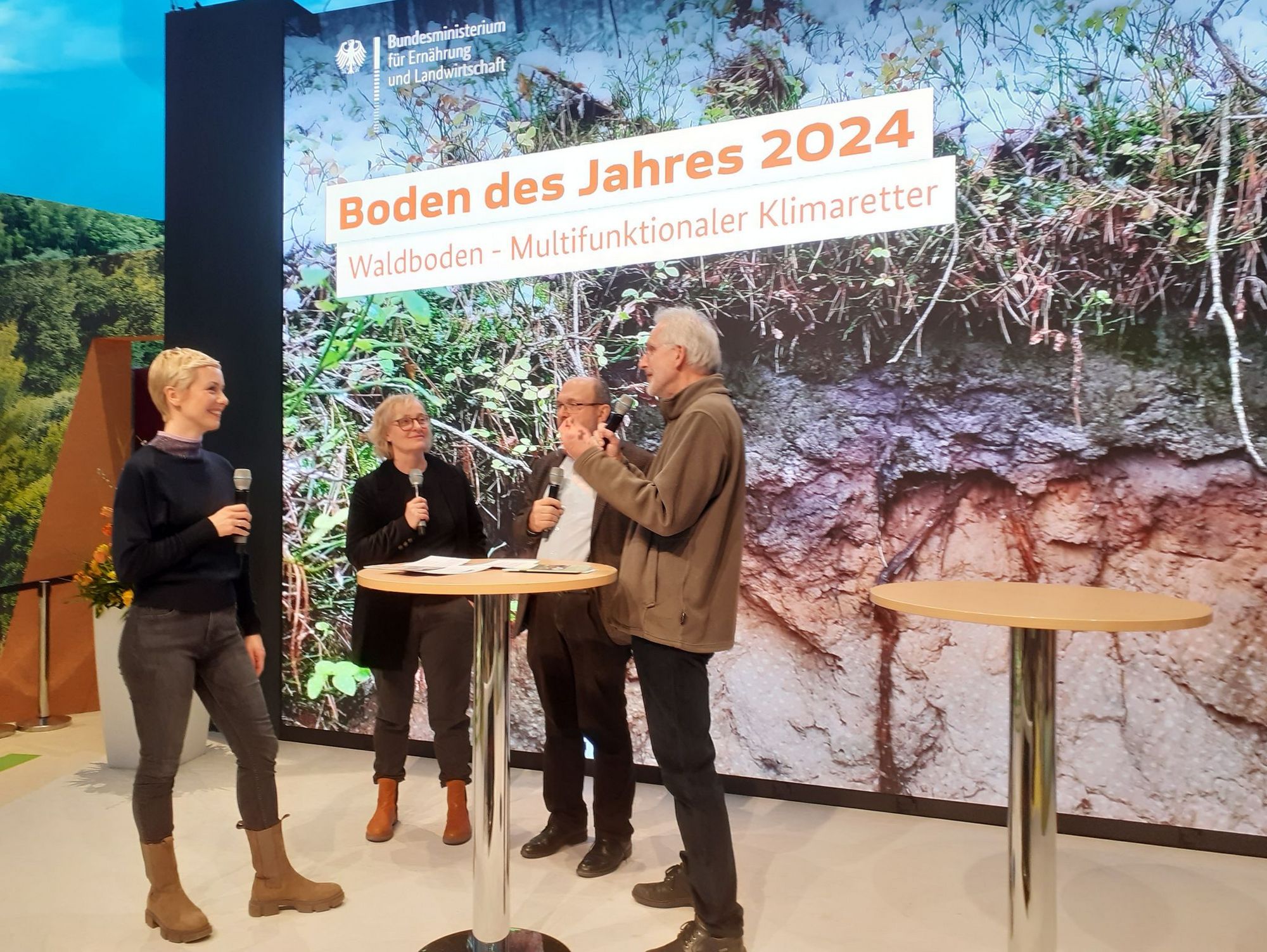 Auf der Bühne in der BMEL-Halle wird der Boden des Jahres 2024, der Waldboden, vorgestellt. Vier Personen im Gespräch.