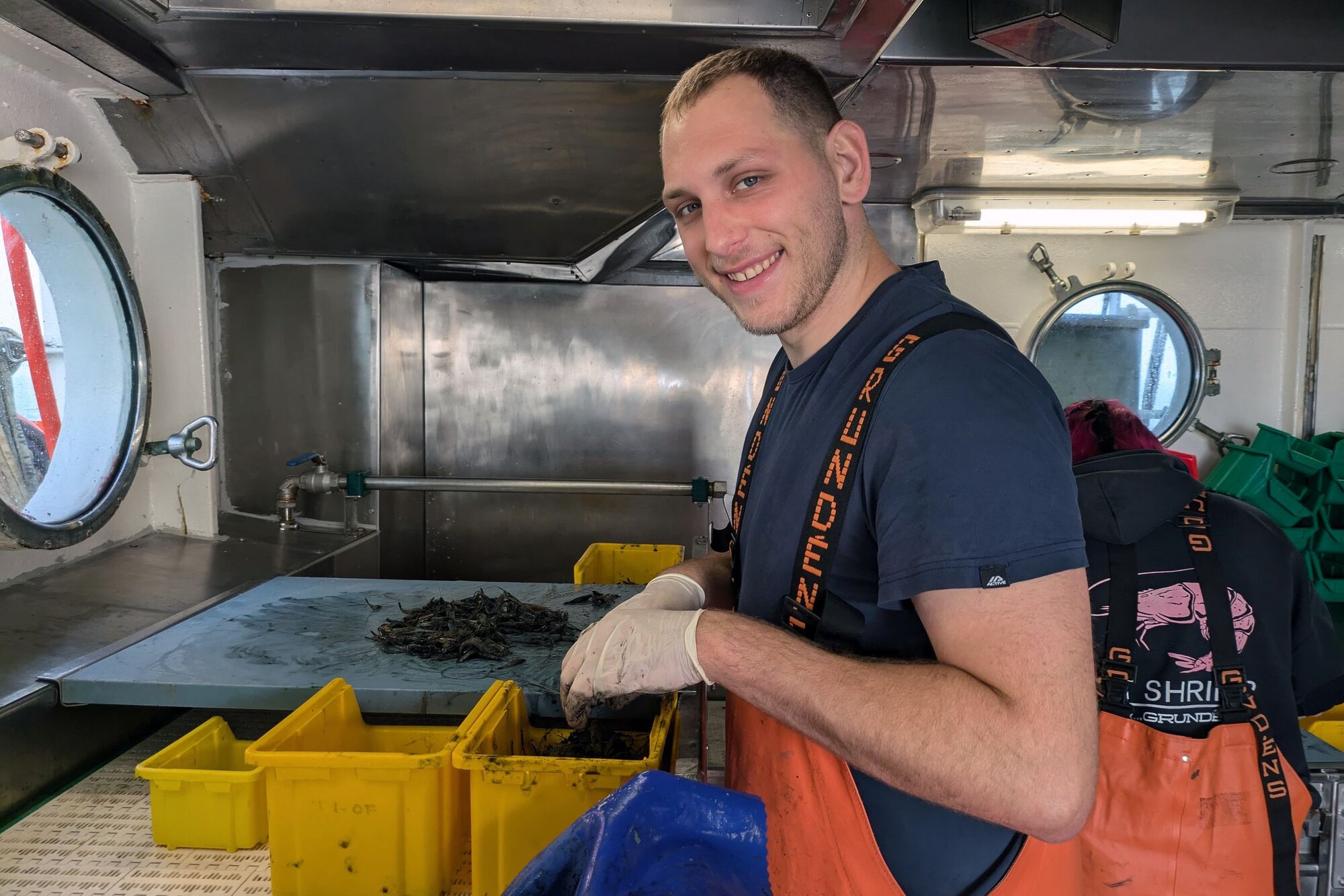 Tim bei der Krabbenfeinsortierung. Ein Mann steht am Förderband und sortiert einen Mix aus Krabben, Quallen, Muscheln und kleinen Fischen.