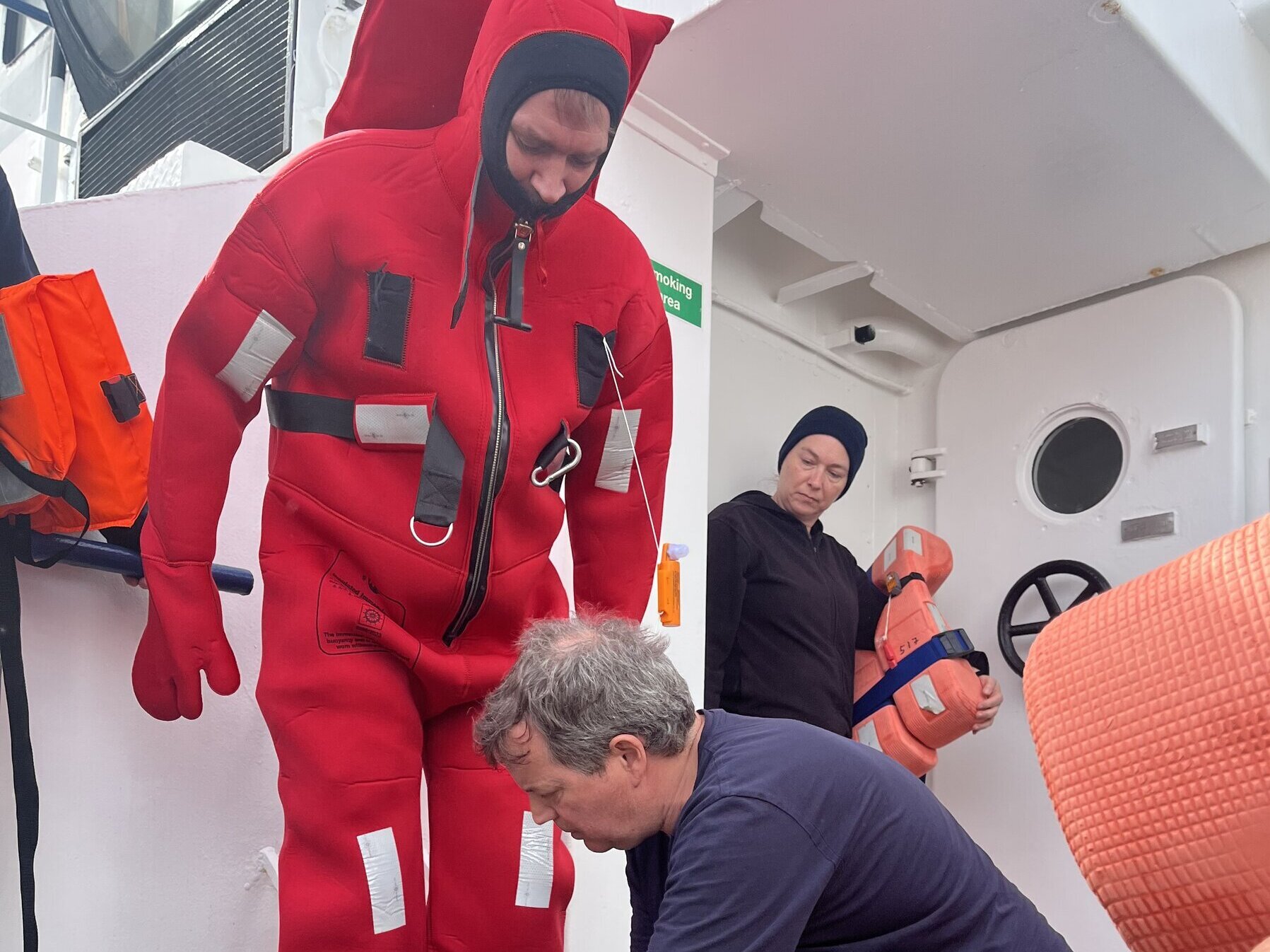 Mehrere Personen an Deck, eine Person steckt in einem roten Seerettungsanzug.