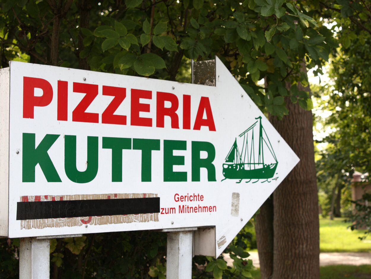 Ein weißes Holzschild, das einen Pfeil nach links darstellt auf dem steht in rot "Pizzeria" und darunter in grün "Kutter" in der Pfeilspitze ist die Zeichnung eines Fischkutters.