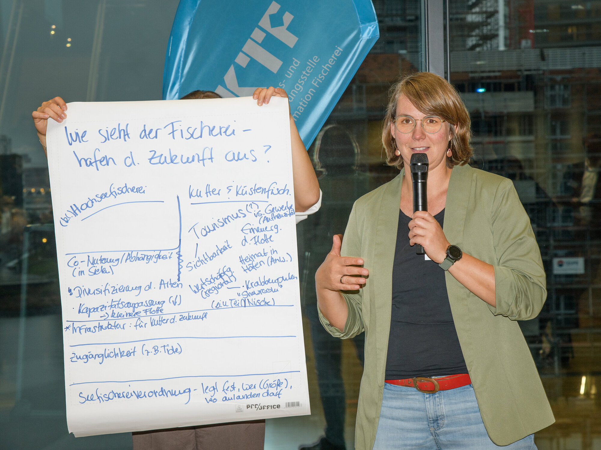 Fanny Barz presents the findings from the dialogue workshop on fishing ports. Portrait of a woman with a poster bearing writing at her side.