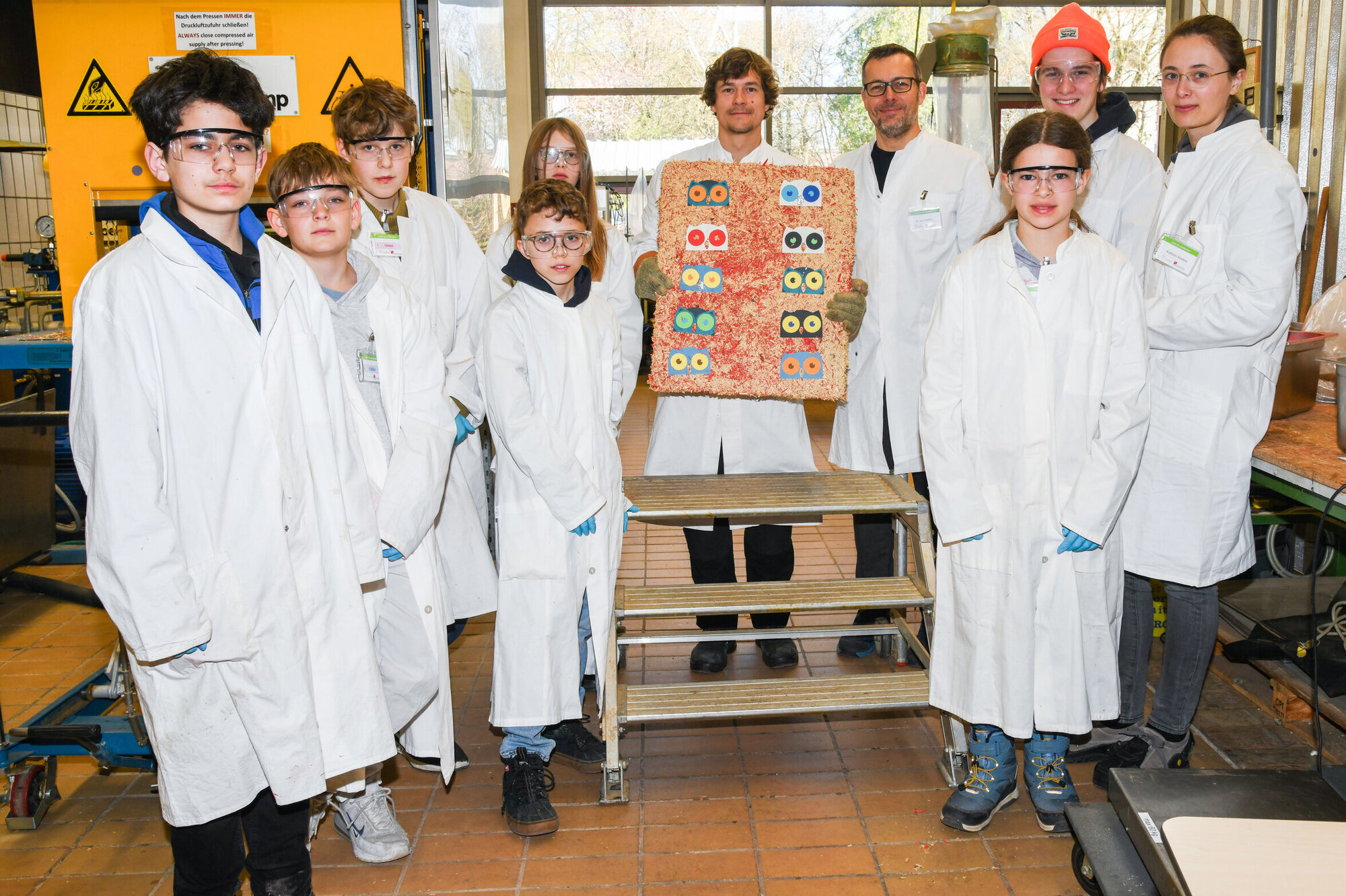 In der Mitte ein erwachsener Mann, der eine Spanplatte hält. Um ihn herum stehen Kinder und Jugendliche. Alle tragen Laborkittel.