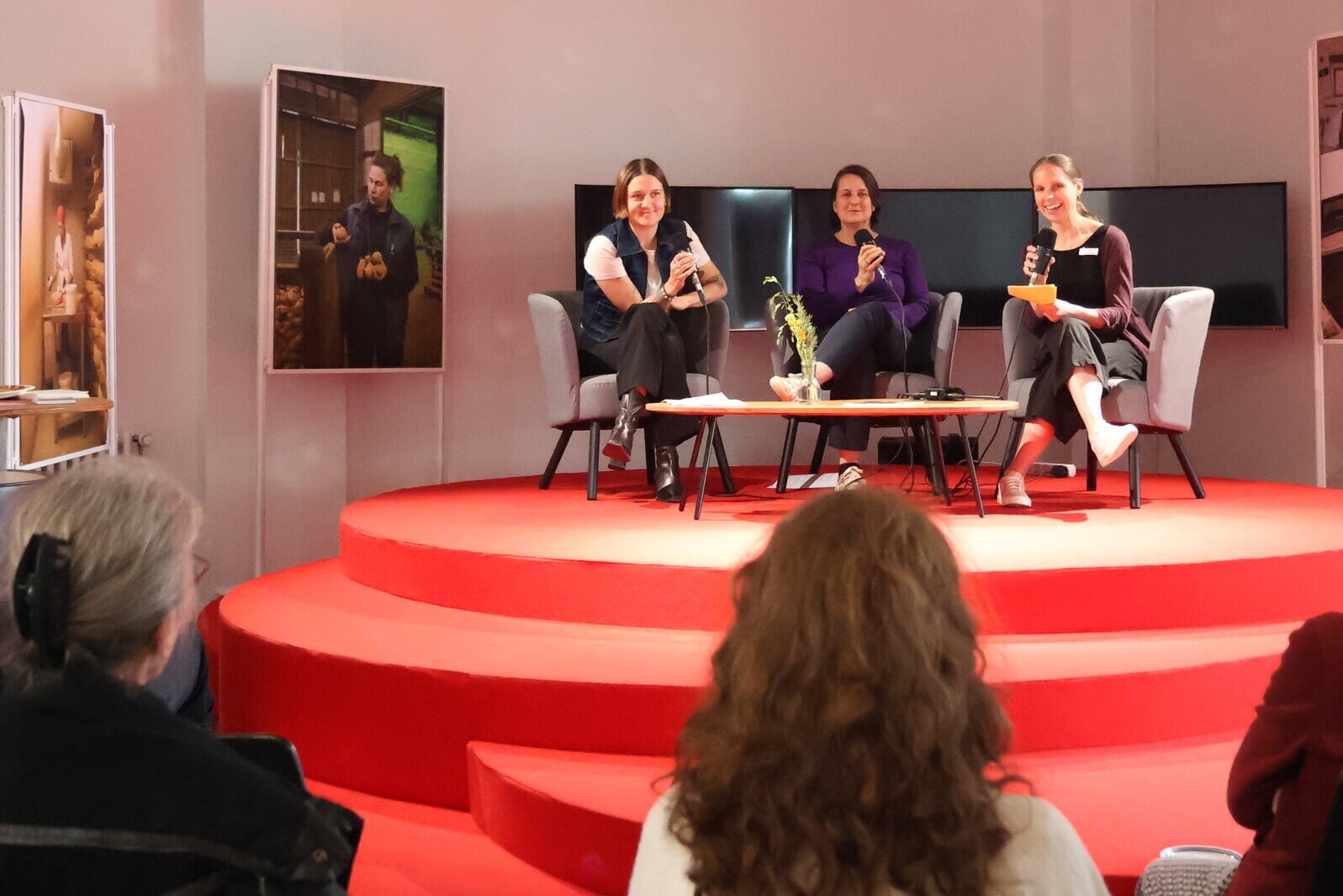 Three women with microphones sit on a red stage.