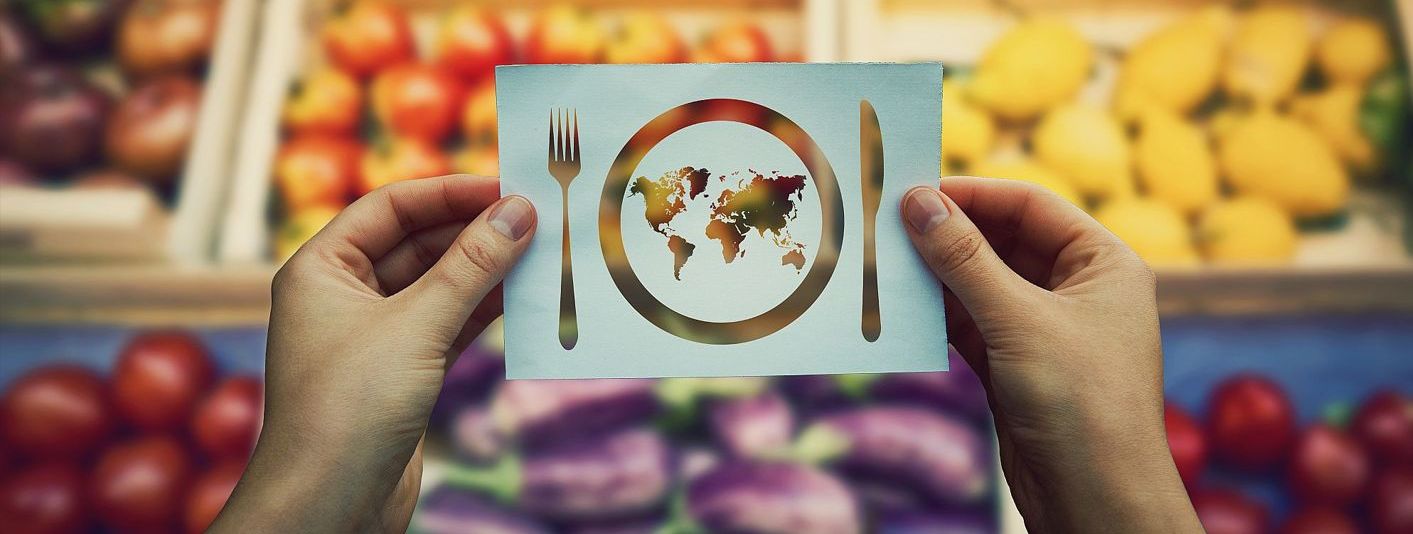 Hände halten ein Blatt Papier, auf dem ein Teller und Besteck zu sehen sind. Auf dem Teller ist eine Weltkarte gezeichnet, im Hintergrund ein Obst- und Gemüseregal im Supermarkt.