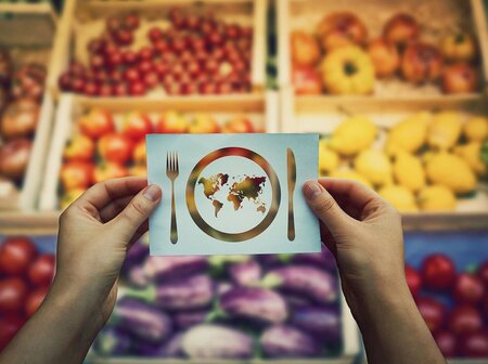 Hände halten ein Blatt Papier, auf dem ein Teller und Besteck zu sehen sind. Auf dem Teller ist eine Weltkarte gezeichnet, im Hintergrund ein Obst- und Gemüseregal im Supermarkt.