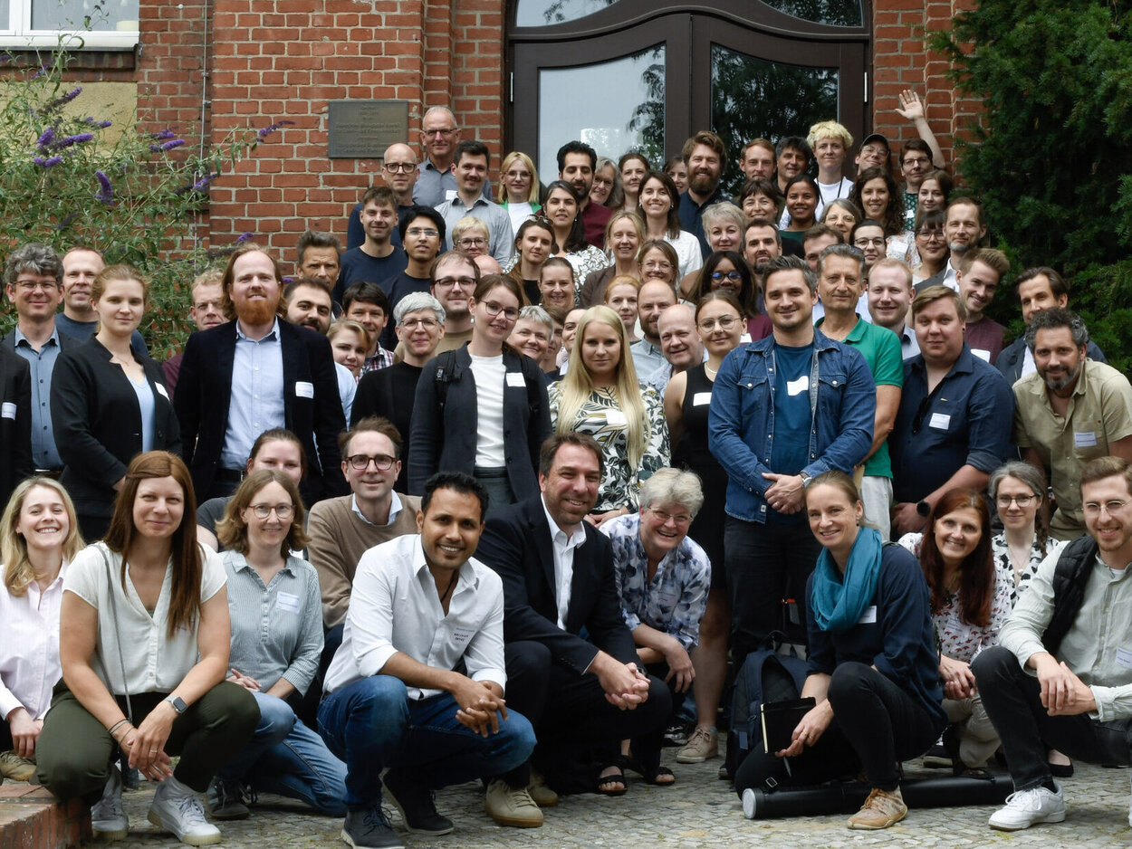 Große Gruppe von etwa 60 Personen vor einem Backsteingebäude, einige sitzen, die meisten stehen, gemischtes Alter und Geschlecht, lächelnd