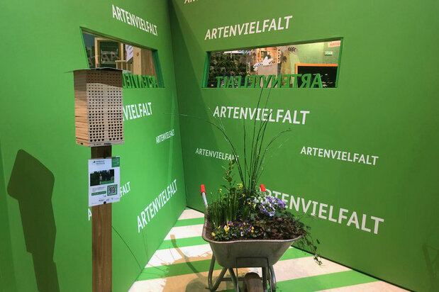 Ein grünes Strukturelement in der Halle mit der Aufschrift Artenvielfalt. Davor eine bepflanzte Schubkarre und eine Wildbienen-Nisthife.