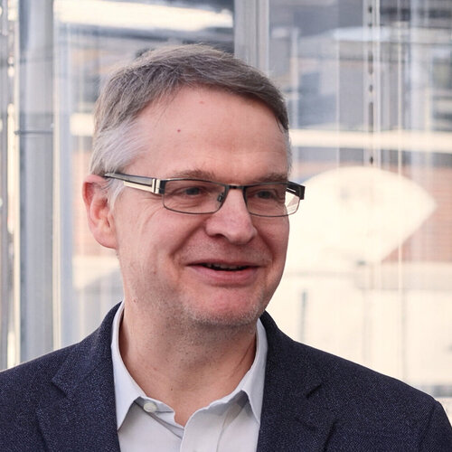 Porträtaufnahmen eines Manns mit Brille und kurzen grauen Haaren. Er steht un gläsernem Gebäude und lächelt.