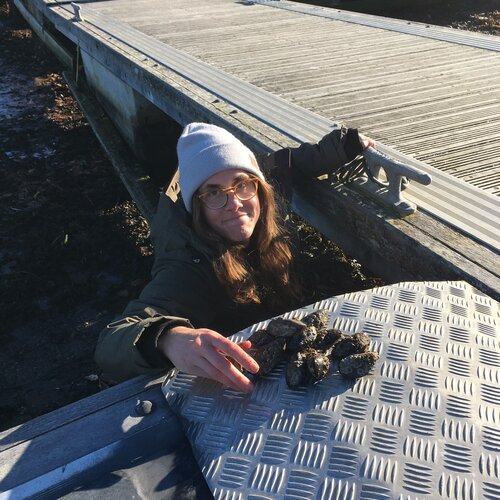 Frau mit Mütze und Jacke neben einem Steg greift nach einer Gruppe Muscheln