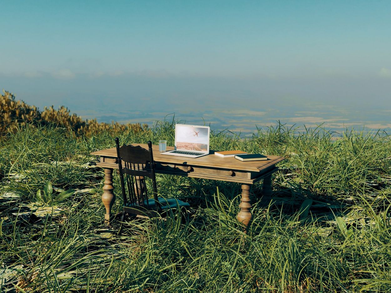 The concept of working from anywhere if you only have your laptop with you. A table and chair are placed in the centre of a field. There is a computer, some books and a glass of water on the table.