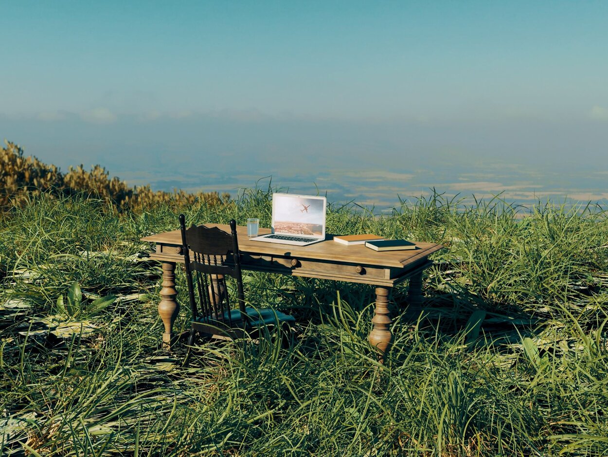 Working from home in a remote location outside The concept of working from anywhere if you only have your laptop with you. A table and chair are placed in the centre of a field. There is a computer, some books and a glass of water on the table.