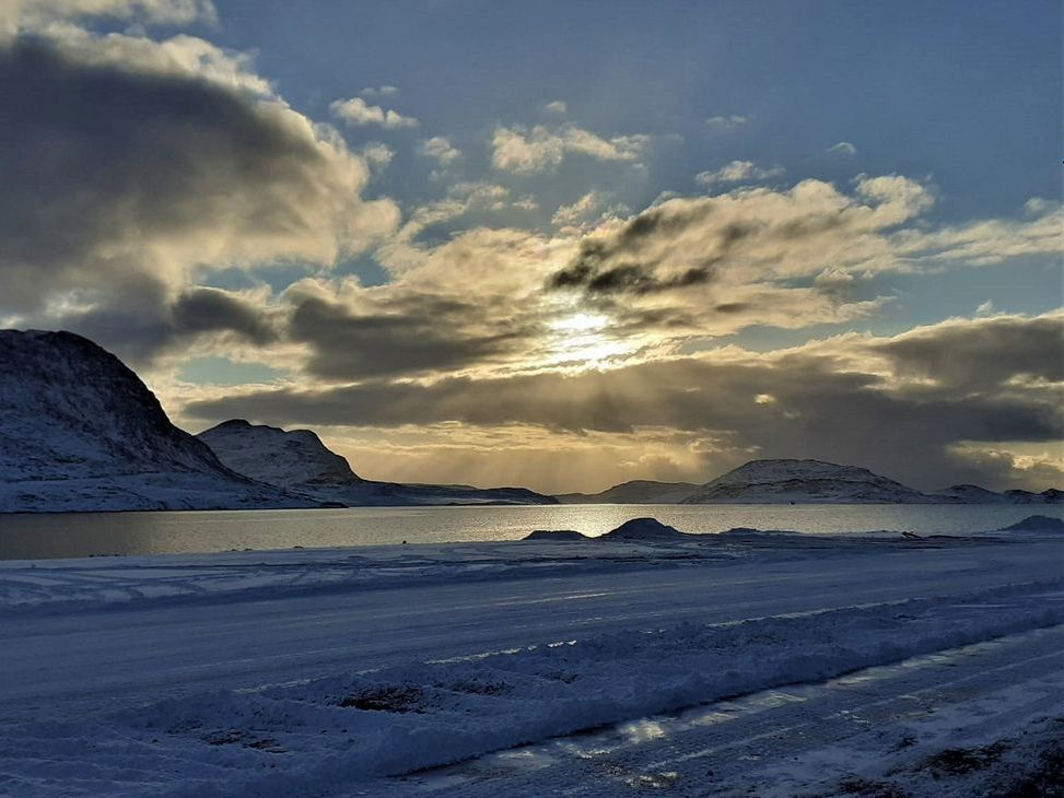 Die Landschaft auf Grönland mit wolkigem Himmel, Sonne, Bergen, Schnee und Wasser.