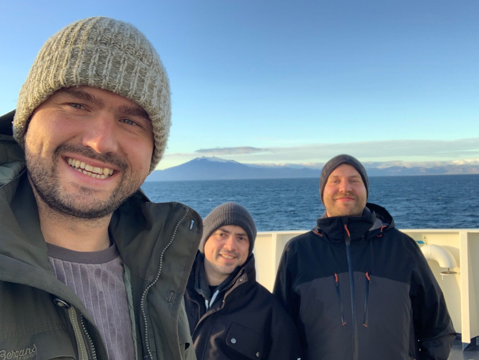 Drei Personen auf einem Selfie - im Hintergrund Meer, blauer Himmel und die Küste Grönlands.