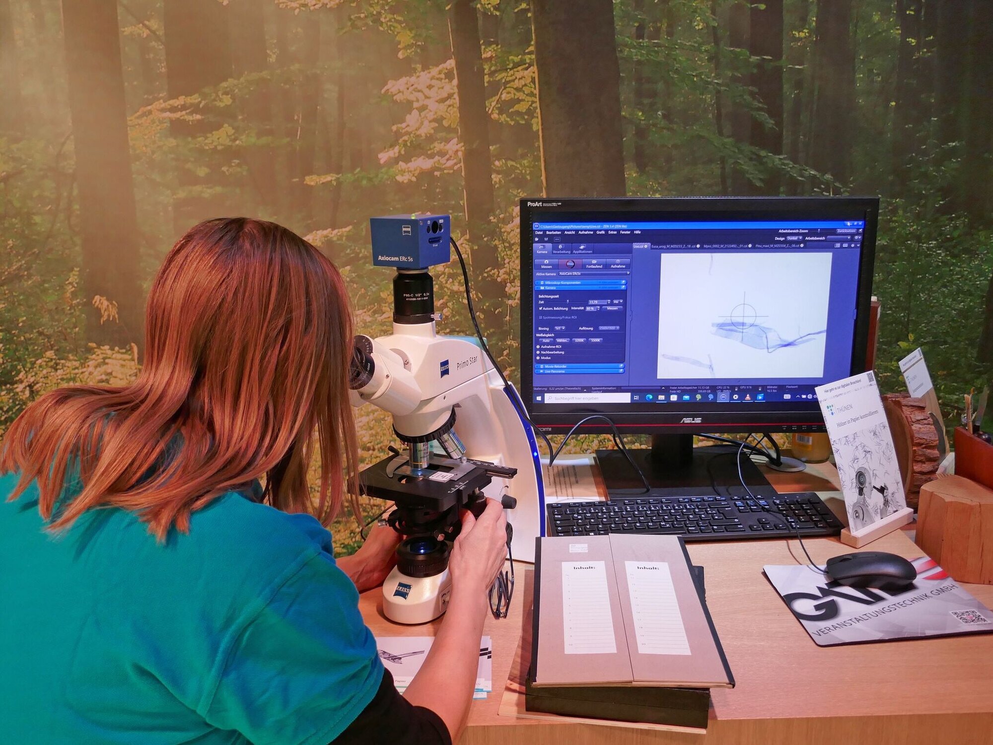 Eine Frau schaut durch ein Mikroskop. Auf einem danebenstehenden Monitor wird das Mikroskopierbild angezeigt.