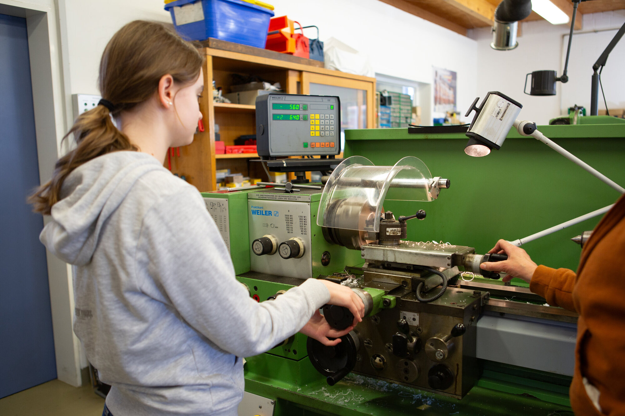 Eine junge Frau bedient eine Drehmaschine in einer Werkstatt. Sie trägt einen grauen Hoodie und konzentriert sich auf die Maschine, während eine andere Person ihr assistiert. Im Hintergrund sind Werkzeuge und Materialien sichtbar.