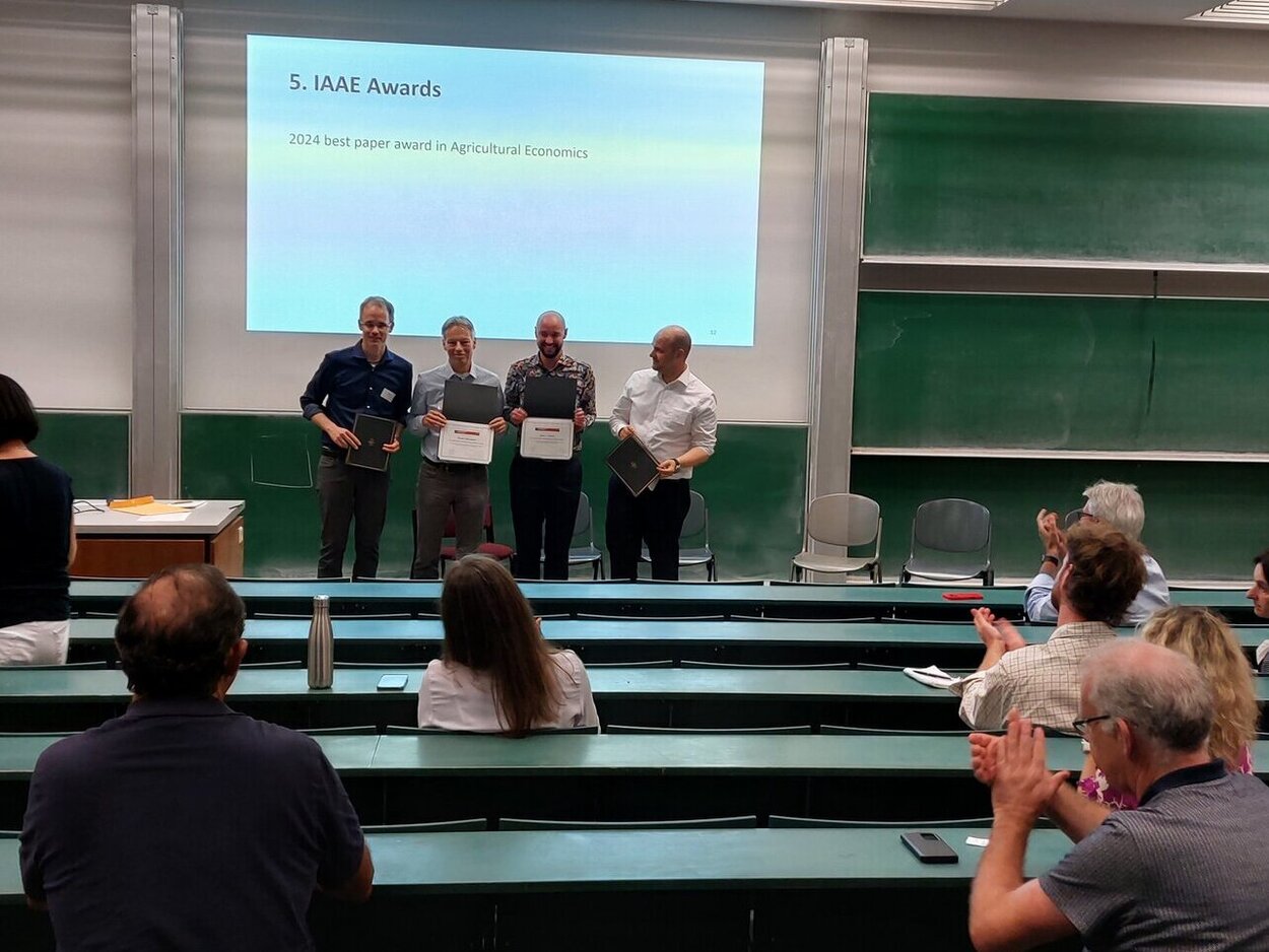Vier Personen stehen vor einer Leinwand in einem Saal und Zuschauer applaudieren.
