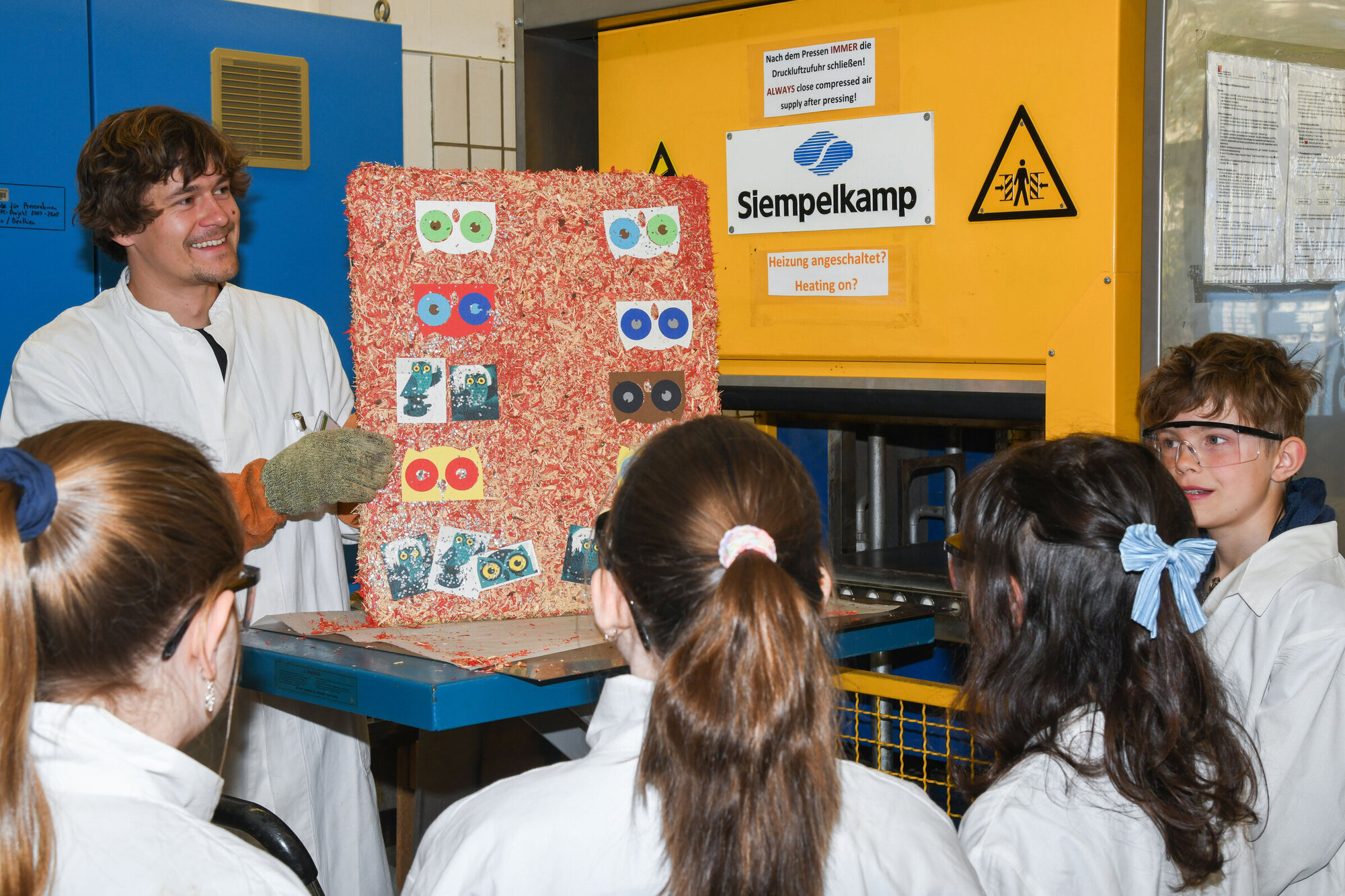 Kinder stehen um einen Mann mit einer Spanplatte herum. Alle tragen Laborkittel.