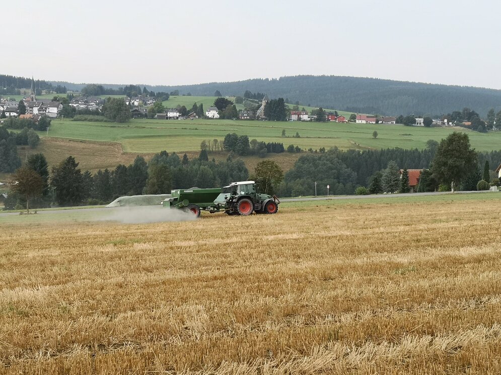 A tractor drives across a harvested grain field in Germany, spreading a white-brown powder.