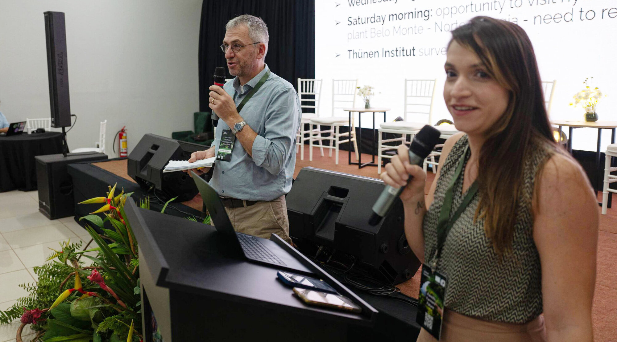 Zwei Personen stehen an einem Rednerpult mit Mikofonen in der Hand und schauen in die Kamera.