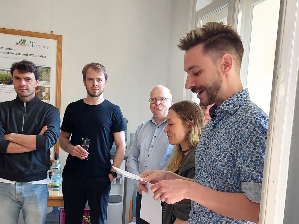 Aufnahme mit vier Personen im Hintergrund. Ein Mann rechts im Bild trägt einen Text vom Blatt vor.