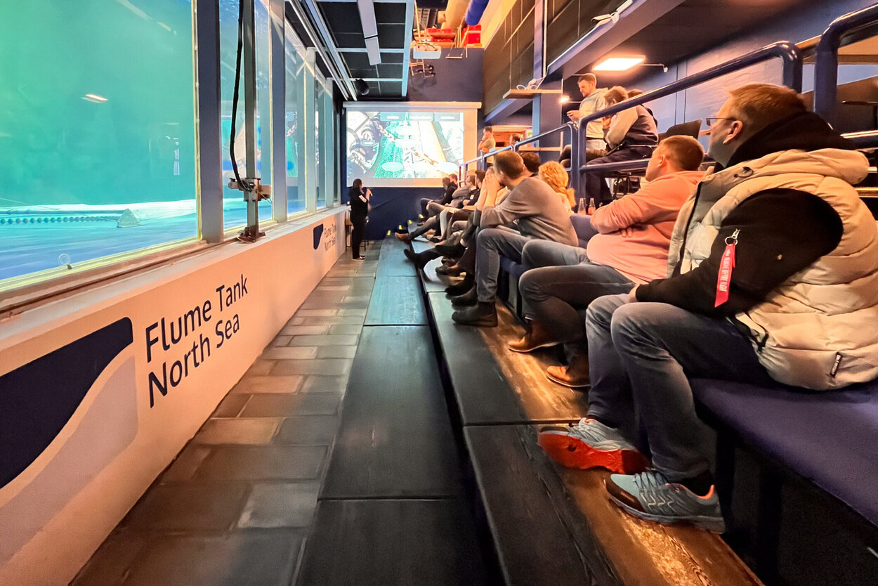 Workshop participants sit in front of the large window and observe a beam trawl model in the water.