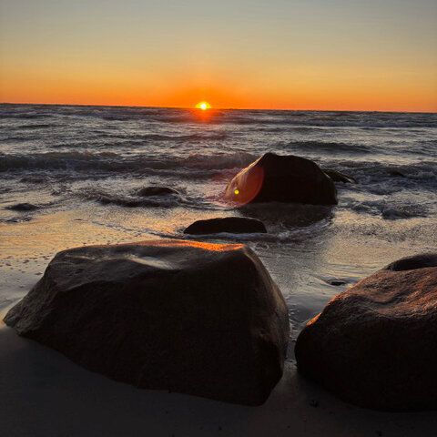 The sun sinks into the sea, with rocks and beach in the foreground.