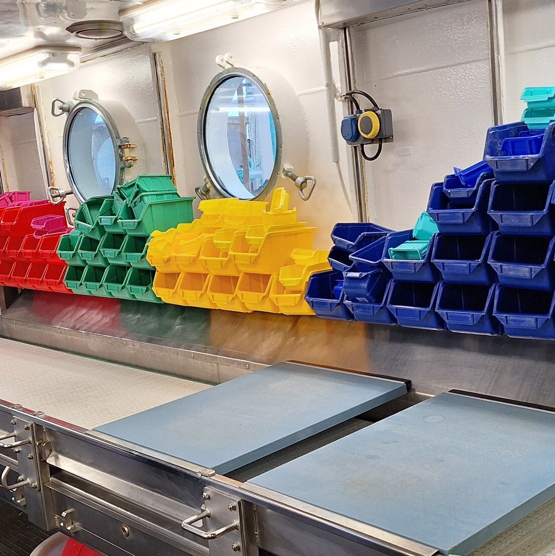 Im Labor des Schiffes hängen über dem Fließband Stapel mit kleinen roten, grünen, gelben und blauen Plastikkiisten.