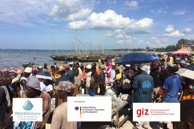 Zahlreiche Menschen auf einem Fischmarkt an einem Strand in Tansania. Im Hintergrund einfach Boote und einige Menschen im Wasser stehend.