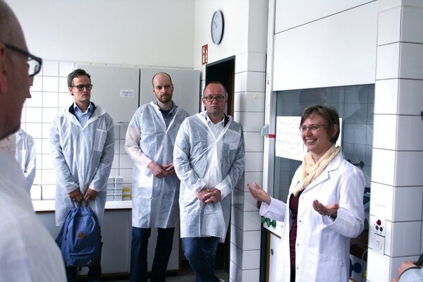 Eine Frau im weißen Kittel steht mit 4 anderen Männern, ebenfalls mit Kittel im Labor und erklärt die Arbeit auf Wurzelebene.