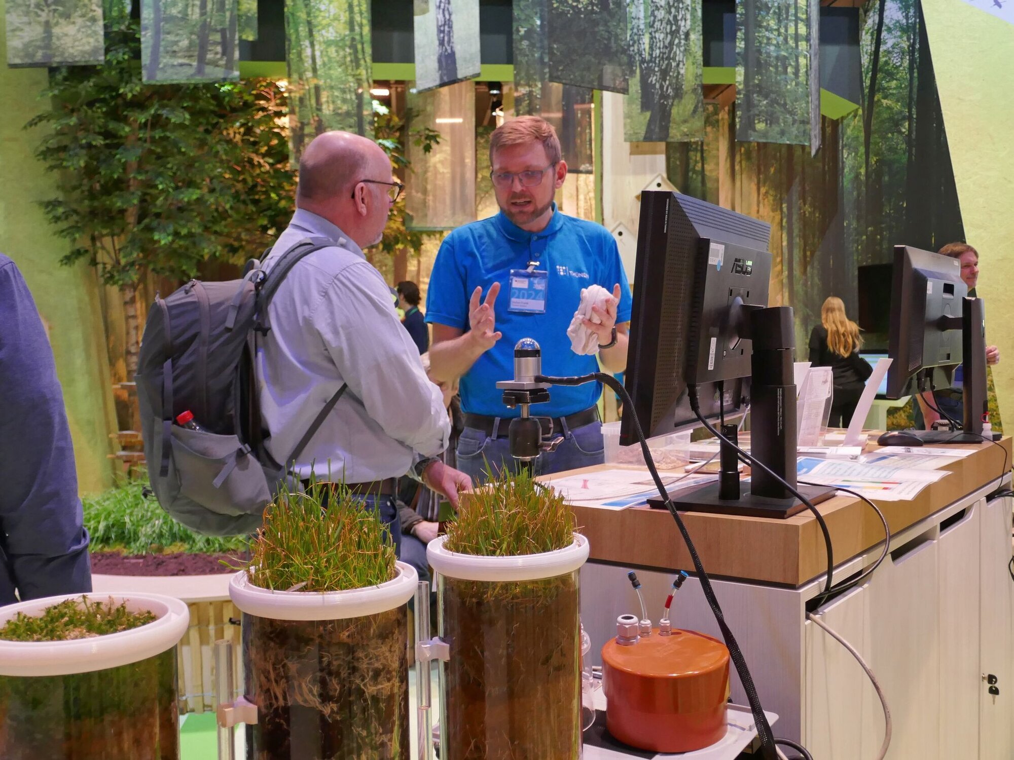 Glayzylinder mit verschiedenen Böden. Im Hintergrund zwei Menschen, die diskutieren.