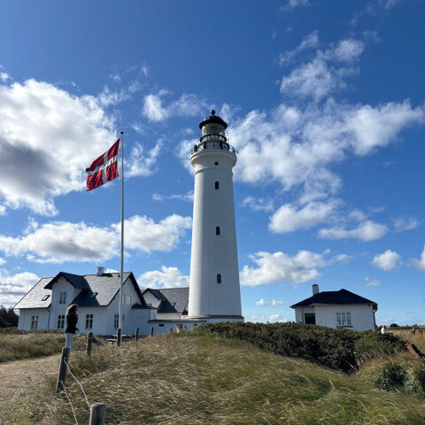 A white lighthouse with two buildings, also white, in front of which the Danish flag is flying.