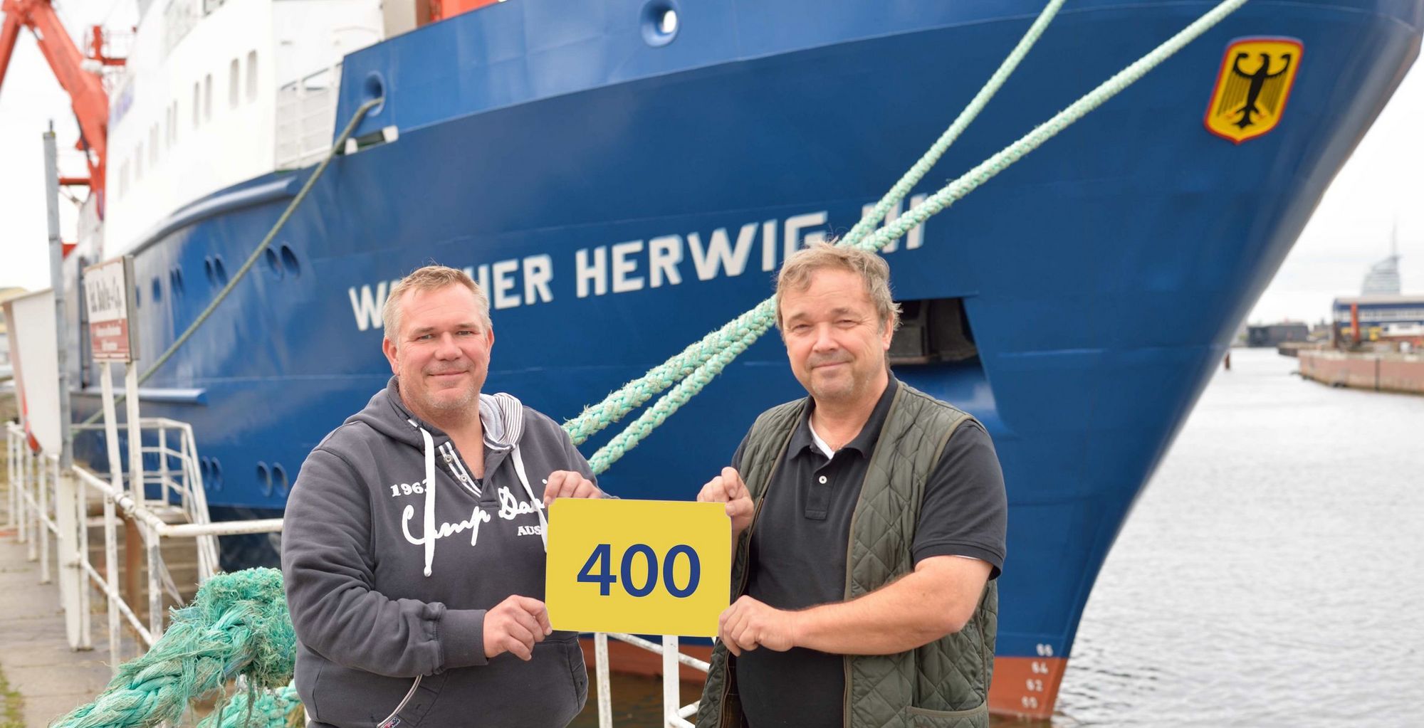 Vor dem Forschungsschiff Walther Herwig III halten der Kapitän und der Fahrtleiter ein Schild mit der Zahl 400 in die Höhe.