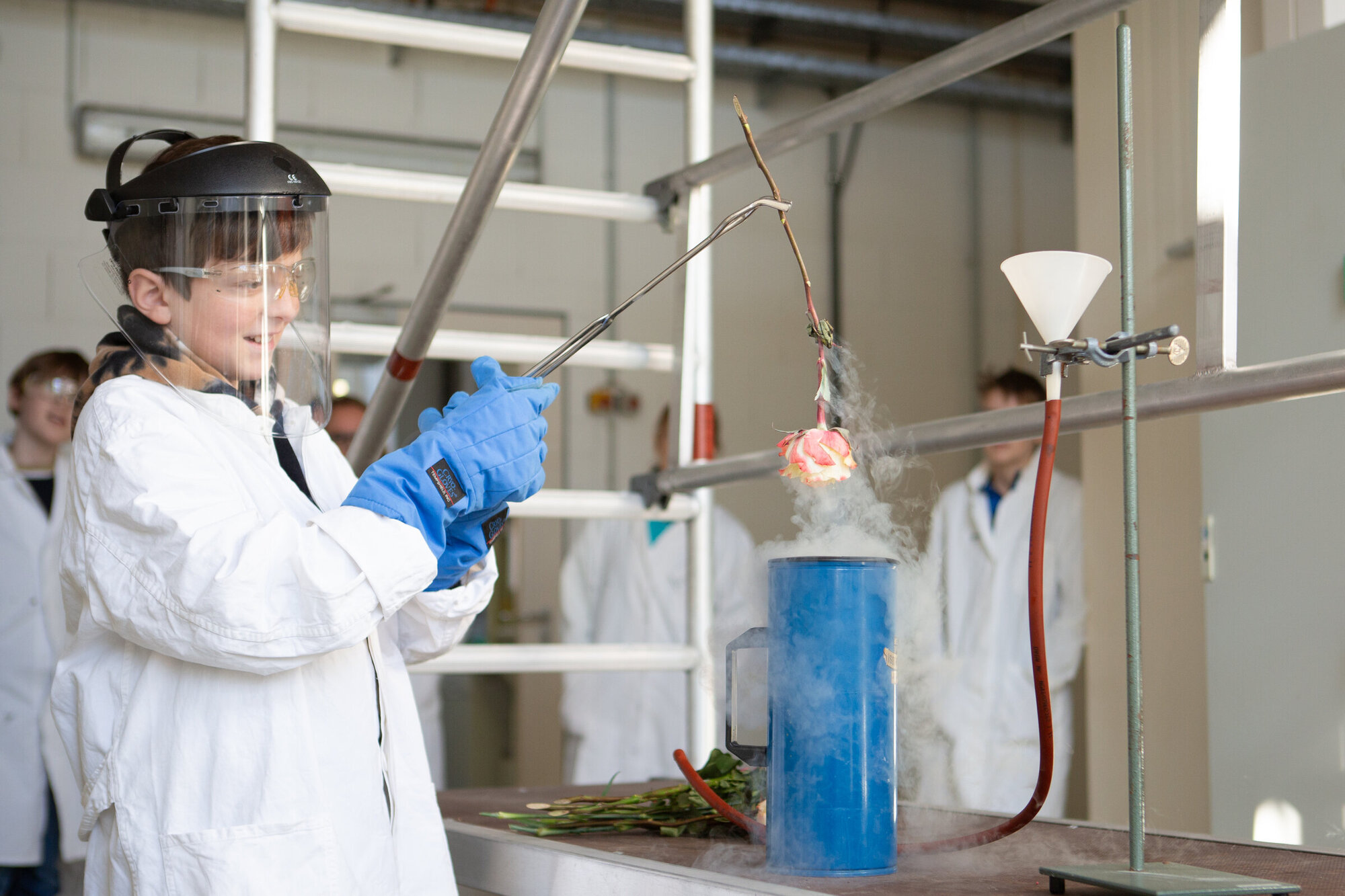 Ein Junge im Laborkittel und Gesichtschutz hält mit einer Zange eine Rose über einem langen, blauen Glasgefäß. Aus dem Gefäß steigt weißer Dampf auf.