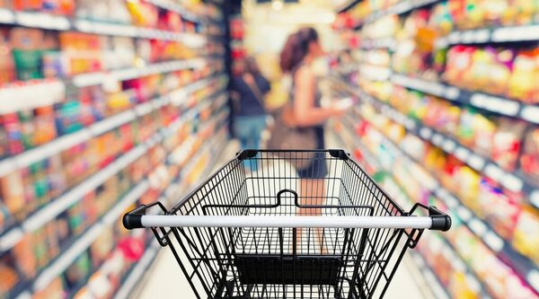 Ein Einkaufswagen steht in einer Supermarktregalreihe, im Hintergrund verschwommen eine Person